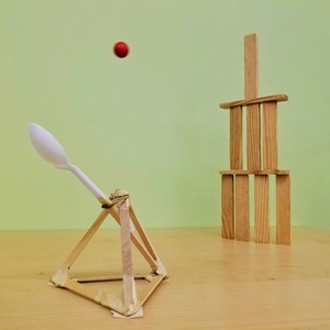 A catapult made of popsicle sticks and a spoon launches a ball at a wooden block tower.