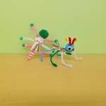 Two Jitterbugs on a table, made of popsicle sticks, pompoms, googly eyes, and pipecleaners. They have small motors connected via wires to coin cell batteries.